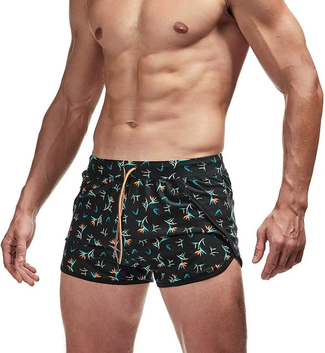 A man wearing black swim trunks with a colorful tropical bird-of-paradise pattern, shown from the chest down on a white background.