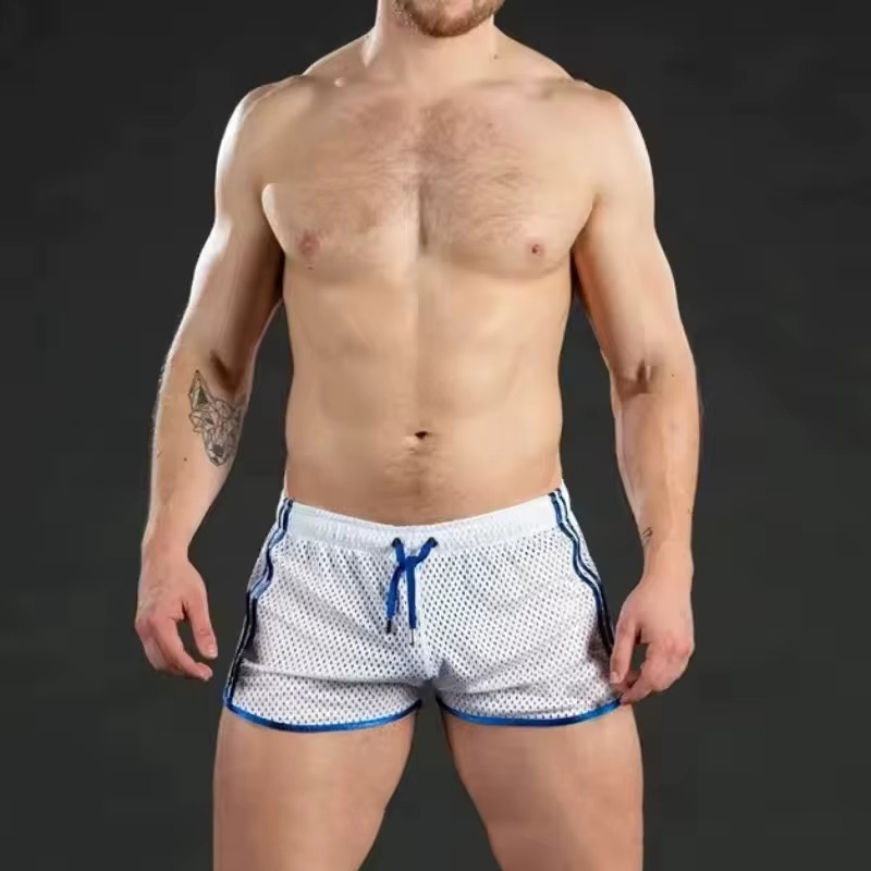 A shirtless male model wearing white mesh athletic shorts with blue trim and a blue drawstring, photographed against a dark gray studio background.